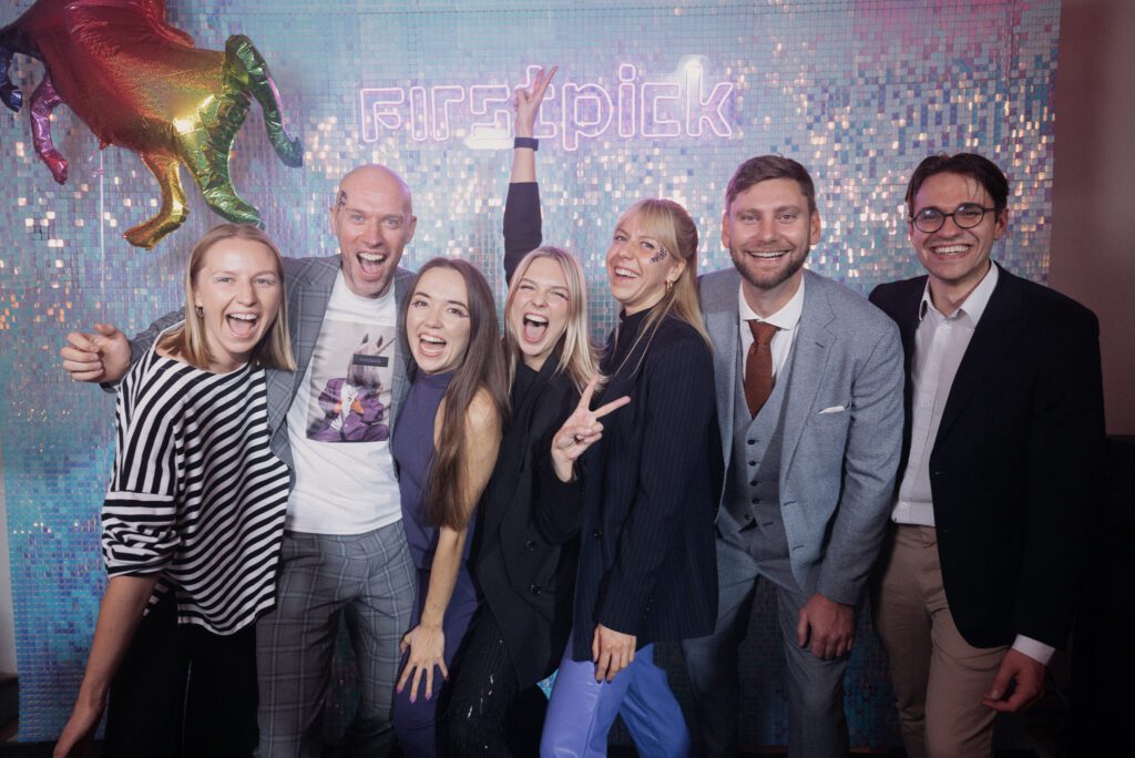 Group of seven people smiling and posing in front of a shimmering backdrop with a neon “firstpick” sign and a colorful unicorn decoration, creating a lively celebration atmosphere.