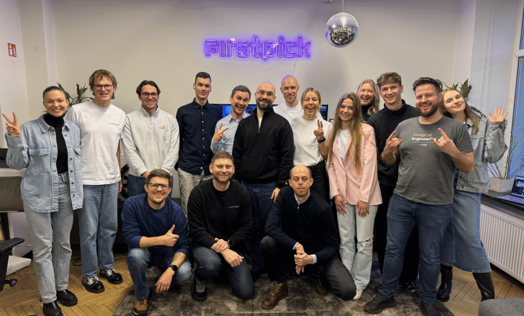 Group photo of the FIRSTPICK team smiling and posing together in an office space. A neon ‘firstpick’ sign and disco ball hang on the wall behind them, creating a cheerful and modern atmosphere. The team is casually dressed, showing peace signs and friendly gestures.