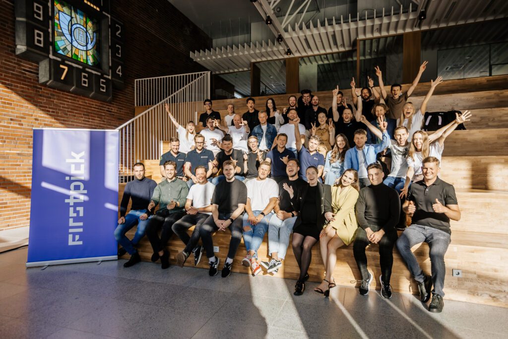 Group photo of FIRSTPICK Accelerator Batch 2 cohort sitting and standing on wooden steps in a bright modern venue. The team and founders are smiling and cheering, with a purple FIRSTPICK banner on the left and sunlight streaming through the windows.
