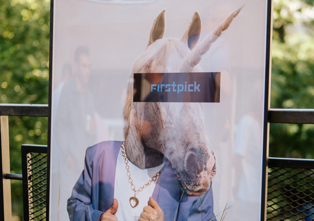 Poster featuring a humorous image of a unicorn in a suit with gold jewelry and a “firstpick” label across its eyes, displayed outdoors surrounded by green plants.
